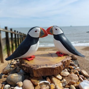 May include: Two carved wooden puffin bird ornaments. They have black and white plumage, red feet, and colourful beaks. The puffins are positioned on a wooden slice, with a beach and sea in the background. The birds are facing each other.