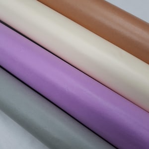 May include: An angled shot of several rolls of paper in different colours. The colours include light grey, lavender, cream, and bronze-brown. The paper appears smooth and tightly rolled. The image is well-lit, showing the subtle variations in the paper's texture.