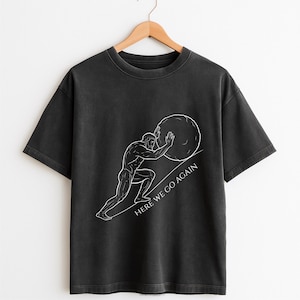 Puede incluir: Camiseta gris oscuro con una ilustraci&oacute;n blanca de un hombre empujando una gran piedra cuesta arriba. El texto "HERE WE GO AGAIN" est&aacute; impreso en blanco. La camiseta est&aacute; colgada en una percha de madera.