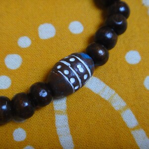 May include: A brown wooden beaded bracelet with a single white and blue patterned bead. The bracelet is on a yellow fabric background with white polka dots.