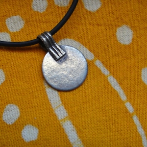 May include: A silver pendant necklace with a round disc charm and a textured surface. The necklace is on a black cord.