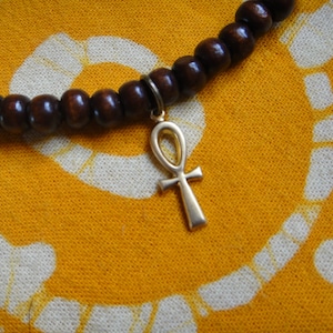 May include: A brown wooden beaded necklace with a gold Ankh pendant. The necklace is on a yellow fabric background with a white pattern.