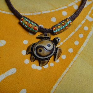 Könnte beinhalten: Eine braune Schnurkette mit zwei grünen, weißen und roten Perlenakzenten. Die Kette hat einen braunen Schildkrötenanhänger mit einem Yin-Yang-Symbol, das in die Schale geschnitzt ist.