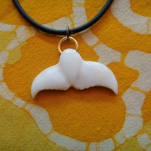 May include: A white whale tail pendant necklace. The pendant is attached to a black cord with a gold-colored ring. The background is a yellow and white patterned fabric. The pendant is a whale tail design.
