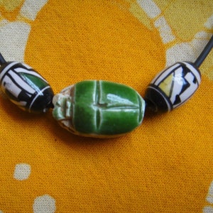 May include: A green ceramic scarab beetle charm on a black cord necklace with white and black patterned beads.