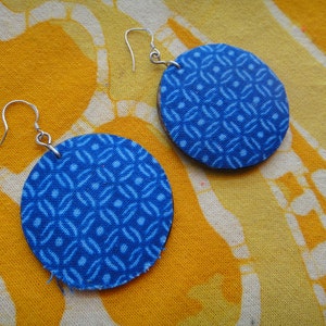May include: A pair of round blue fabric earrings with a geometric pattern. The earrings are suspended from silver-colored ear wires. The earrings are displayed on a yellow and white patterned fabric.