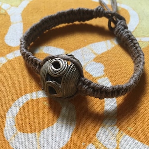 May include: A brown braided bracelet with a silver metal bead featuring a circular design.