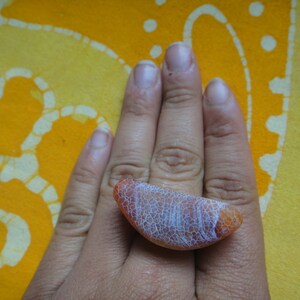 以下が含まれることがあります： 白とオレンジのアゲートのリングで、白いひび割れた模様があります。リングは指にはめられています。