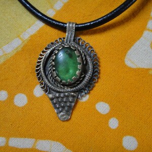 Puede incluir: Un collar con un colgante de piedra verde y un diseño de filigrana de plata. El collar está en un cordón de cuero negro.