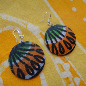 May include: A pair of round earrings with a colourful fabric design. The fabric is orange, black, green and white. The earrings are made with silver hooks.