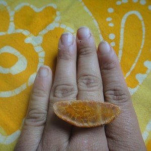 May include: A hand wearing a large, orange agate ring with a unique, natural pattern. The ring is on the index finger and the hand is resting on a yellow and white patterned fabric.