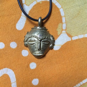 May include: A gold-colored pendant necklace featuring a detailed, stylized face mask with intricate designs. The mask has a raised, textured surface and is suspended from a black cord.