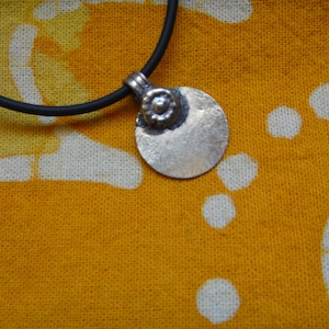 Puede incluir: Un collar de plata con un colgante redondo y un pequeño diseño de flor. El collar está en un cordón negro.