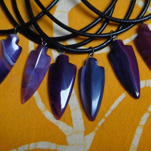 May include: Six purple and white agate arrowhead pendants on black leather cords. The arrowheads are all different shapes and sizes.