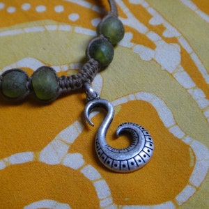 May include: A brown cord necklace with green glass beads and a silver pendant shaped like a hook with a swirl design.