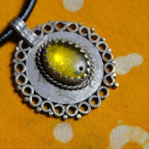 Puede incluir: Un collar con un colgante de plata con una piedra amarilla engastada en un marco decorativo de plata. El colgante está unido a un cordón de cuero negro.