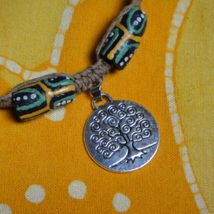 May include: A brown hemp cord necklace with two green, black, and yellow patterned beads and a silver pendant featuring a tree of life design.