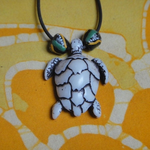 Op de afbeelding: Een ketting met een witte keramische schildpad hanger met zwarte en groene kralen. De schildpad heeft een gedetailleerde schild en hangt aan een zwart koord.