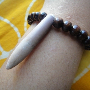 Könnte beinhalten: Ein braunes Holzperlenarmband mit einem langen, hellbraunen Holzanhänger. Das Armband wird am Handgelenk einer Person getragen.