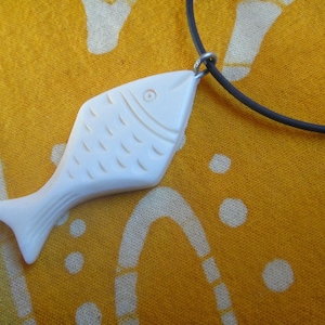 Op de afbeelding: Witte gegraveerde vis-hanger aan een zwarte koordsnoer. De hanger is gedetailleerd met lijnen en een klein oog.
