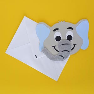 Könnte beinhalten: Ein weißer Umschlag mit einem Cartoon-Elefantenkopf auf gelbem Hintergrund. Der Elefant hat graue Haut, blaue Ohren, einen grauen Rüssel und schwarze Augen. Der Elefant lächelt. Der Umschlag ist geschlossen.