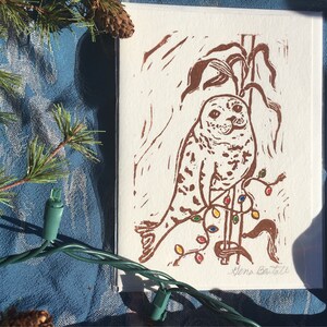 Puede incluir: Una impresi&oacute;n en lin&oacute;leo marr&oacute;n de una foca sentada en una rama con luces de Navidad de colores. La impresi&oacute;n est&aacute; firmada "Elena Battell" en la esquina inferior derecha.