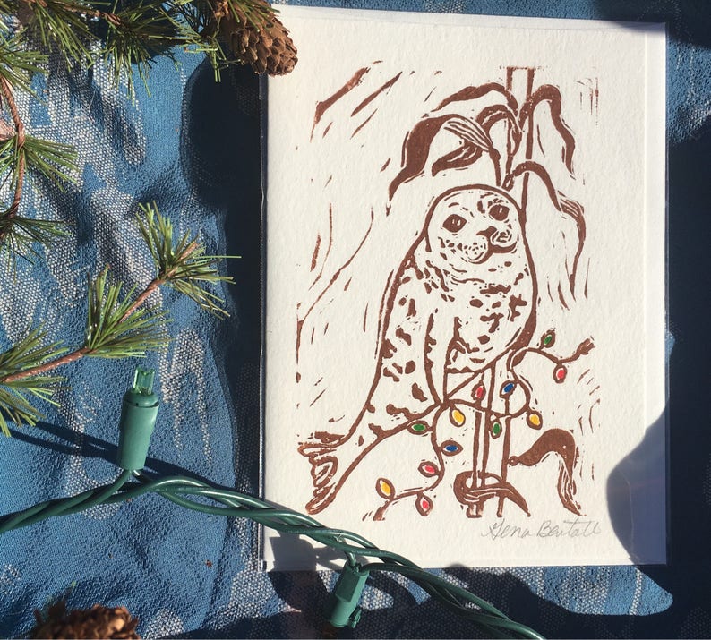 Puede incluir: Una impresi&oacute;n en lin&oacute;leo marr&oacute;n de una foca sentada en una rama con luces de Navidad de colores. La impresi&oacute;n est&aacute; firmada "Elena Battell" en la esquina inferior derecha.