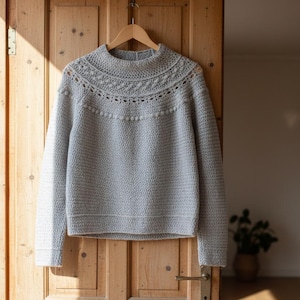 May include: A light gray, hand-crocheted sweater with a high neckline and long sleeves hangs on a wooden hanger. The sweater features a textured stitch pattern and decorative detailing around the neckline. The background is a wooden door.