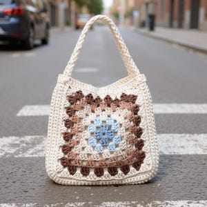 Peut inclure: Sac fourre-tout en crochet crème avec un motif carré multicolore sur le devant. Le motif présente un carré bleu central entouré de carrés marron et beige. Le sac a une poignée tressée et est posé sur un passage piéton.