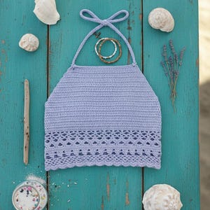 Puede incluir: Un top halter de ganchillo lila con un borde inferior decorativo, presentado sobre una superficie de madera turquesa. La imagen incluye conchas marinas, un pequeño tarro de cristal con elementos decorativos y una ramita de lavanda.