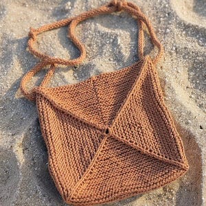 Könnte beinhalten: Eine gestrickte Umhängetasche in einem warmen Braunton, die auf Sand liegt. Die Tasche hat ein strukturiertes Strickmuster und einen langen, geknoteten Riemen. Das Design der Tasche ist einfach und geometrisch, mit einem zentralen Punkt, an dem sich die Strickpaneele treffen.
