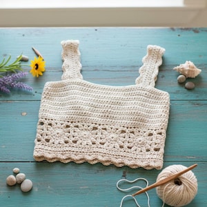 May include: A cream-colored, handmade crochet top with scalloped edges and straps. The top features a floral pattern along the bottom. The top is displayed on a turquoise surface with a ball of yarn and a crochet hook.