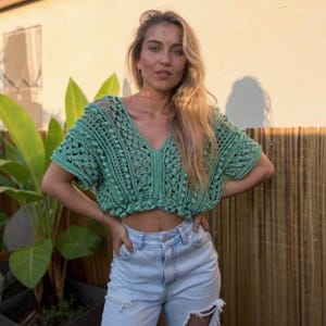 Puede incluir: Top corto de ganchillo verde mar con mangas cortas y cuello en V. El top tiene un diseño texturizado y calado. La modelo lleva vaqueros de mezclilla azul claro. El fondo incluye una valla de bambú y exuberantes plantas verdes.
