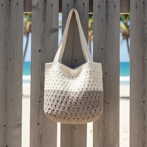 May include: A two-tone crochet tote bag with a cream-colored top and a taupe-colored bottom, hanging on a weathered wooden fence. The bag has a rectangular shape with a wide opening and two cream-colored handles. The background shows a beach scene.