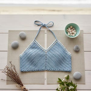 Peut inclure: Haut de bikini en crochet bleu clair avec une bordure blanche, présenté sur un set de table neutre. Le haut fait main présente un motif en chevrons. Des pierres, un petit bol de perles et un bouquet de brindilles complètent la scène.