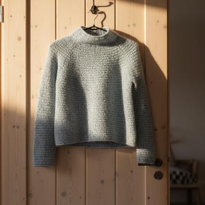 Peut inclure: Un pull en tricot gris clair et épais avec un col montant côtelé est suspendu à un crochet en métal. Le pull a des manches longues et un aspect texturé. Il est présenté contre une porte en bois de couleur claire.