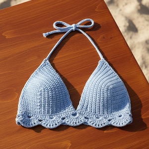 Könnte beinhalten: Hellblaues Häkel-Bikinioberteil mit gewelltem Rand. Die Dreiecks-Cups sind durch eine Nackenbindung verbunden. Das Oberteil wird auf einer Holzoberfläche mit einem Sandstrand im Hintergrund präsentiert.