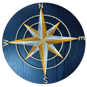 Può includere: Un'applique da parete rotonda in legno blu scuro con un design a bussola in filo d'oro, bianco e giallo. La rosa dei venti ha otto punte, con i punti cardinali etichettati. Il design è centrato sul cerchio di legno.