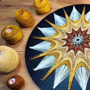 Könnte beinhalten: Ein handgefertigtes String-Art-Stück mit einem Sternmuster in Gelb-, Braun- und Weißtönen auf einer schwarzen, kreisförmigen Basis. Mehrere Spulen gelber und brauner Fäden sind um das Kunstwerk herum verstreut.
