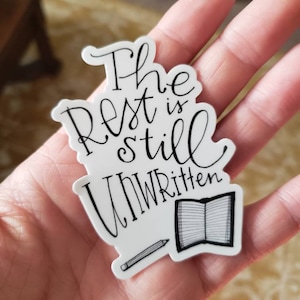 Könnte beinhalten: Weißer Aufkleber mit schwarzem Schriftzug "The Rest is still Unwritten". Der Aufkleber zeigt auch eine kleine Illustration eines offenen Buches und eines Bleistifts. Der Aufkleber wird in einer Hand gehalten.