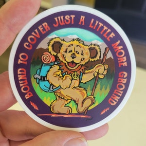 May include: A round sticker with a purple background and a cartoon bear wearing a backpack and holding a walking stick. The text around the bear reads "Bound to cover just a little more ground".