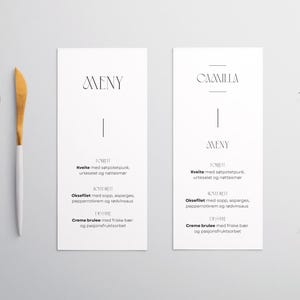 May include: A minimalist dining scene featuring two white-handled utensils with gold-colored heads, two menu cards, and a sprig of greenery. The menu cards have the word "AVENY" and "CAMILLA" printed on them.