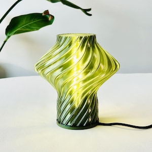 Könnte beinhalten: Eine grüne Tischlampe aus Glas mit einem einzigartigen, gewirbelten Design. Die Lampe hat eine abgerundete Ober- und Unterseite mit einem strukturierten, gerippten Muster. Die Lampe ist beleuchtet und strahlt ein warmes Licht aus. Ein schwarzes Netzkabel erstreckt sich von der Basis.