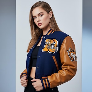 May include: A navy blue and tan varsity jacket with orange and white accents. The jacket features a large orange letter "A" with a football helmet patch and a wildcat patch. The sleeves are tan, and the jacket has a button-up front.