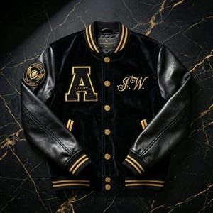 Könnte beinhalten: Schwarze Collegejacke mit goldenen Akzenten. Mit einem großen goldenen "A LUXURY" Aufnäher, goldenen Verzierungen und Lederärmeln. Enthält einen "LONDON EST. 2023" Aufnäher und die Initialen "J.W." in goldener Schrift. Die Jacke hat eine Knopfleiste.