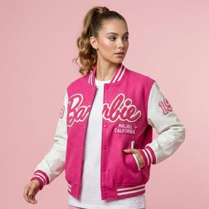 Könnte beinhalten: Eine pinkfarbene und weiße Collegejacke mit dem Wort "Barbie" in Schreibschrift auf der Brust. Die Jacke hat weiße Ärmel und den Text "Malibu California" unter dem Barbie-Logo. Die Zahl "19" befindet sich auf dem rechten Ärmel.