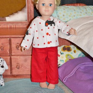 May include: A blonde doll wearing a white and red pajama top with a black dog graphic and red pants. The doll is standing in front of a wooden dresser and a bed with a purple blanket and a patterned pillow.