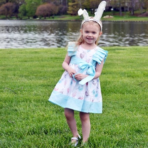 May include: A young girl wearing a light blue dress with a white and pink floral pattern and a white bunny ear headband. She is standing in a grassy field with a lake in the background.