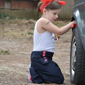 May include: A young person wearing a white tank top and a blue skirt with red buttons is kneeling next to a car. They are holding a screwdriver and have a red bandana tied around their head.