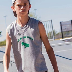 May include: A young person wearing a grey sleeveless shirt with the text "I do my own stunts" and a green skateboard graphic. They are also wearing a grey baseball cap and a silver necklace.